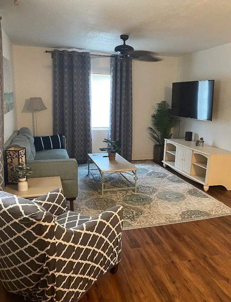Living room with a teal sofa, a patterned armchair, a wall-mounted television and a rug over wood floors