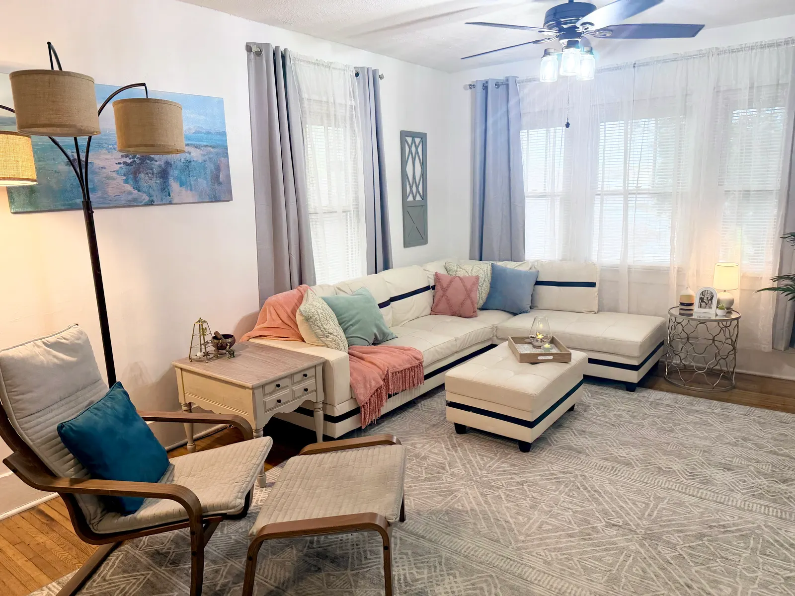 Living room with a white sectional sofa and a matching ottoman on a patterned rug, a wooden armchair and footstool, a floor lamp, and curtained windows along two walls, under a ceiling fan