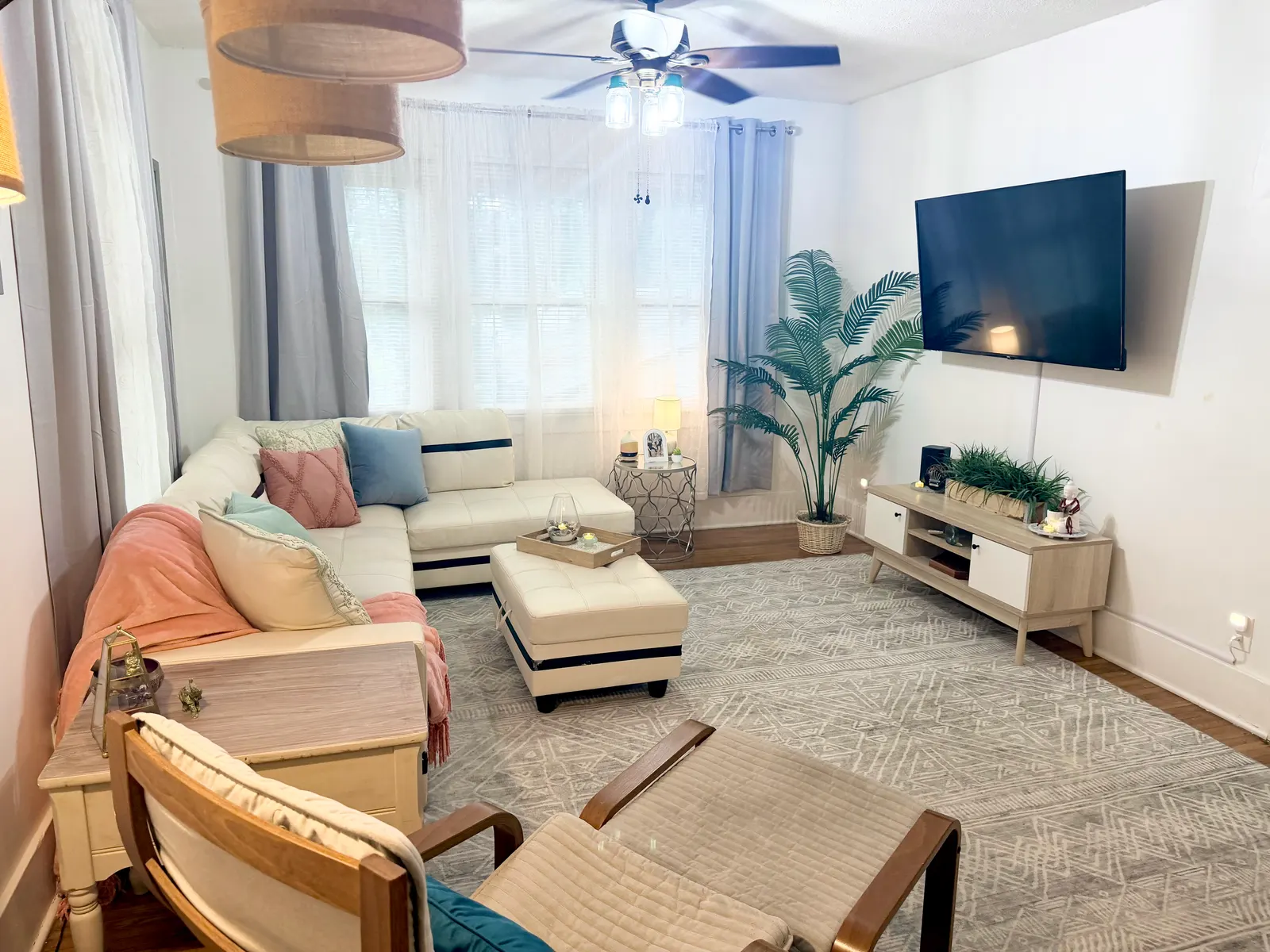 The same living room from the other end, with a television mounted on the wall above a low media unit, a tall potted palm in the corner, and the sectional beneath the ceiling fan