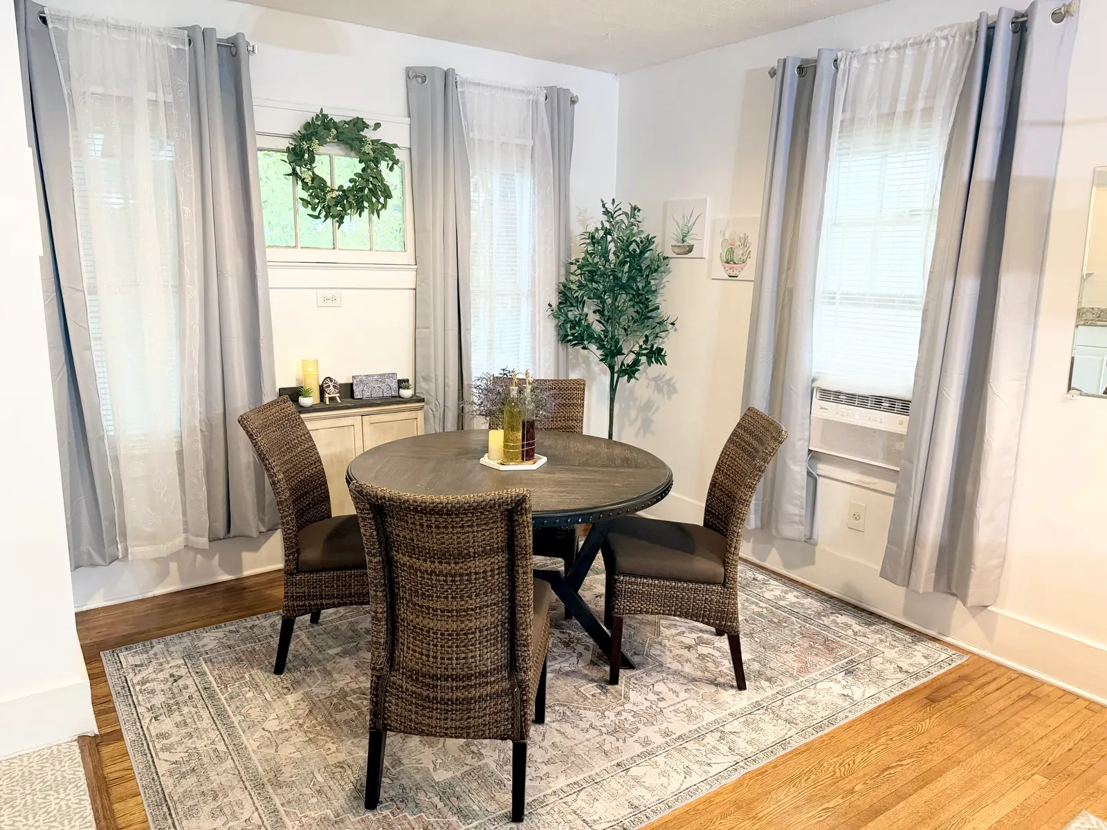 Dining area with a round table and four woven-back chairs on a rug over hardwood, a sideboard against the wall, and a window air-conditioning unit in one of three curtained windows
