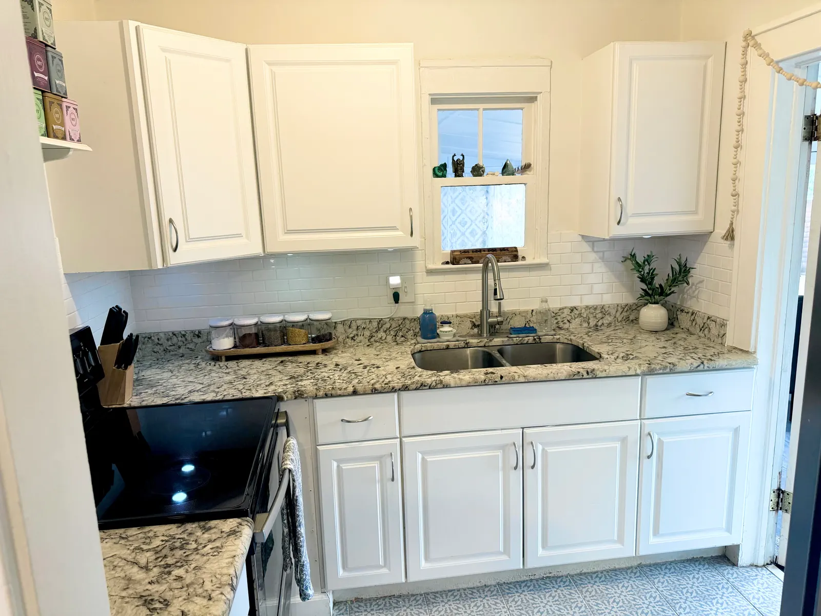 Kitchen with white cabinets, granite counters, a subway-tile backsplash, a two-bowl stainless sink under a window, and a patterned tile floor