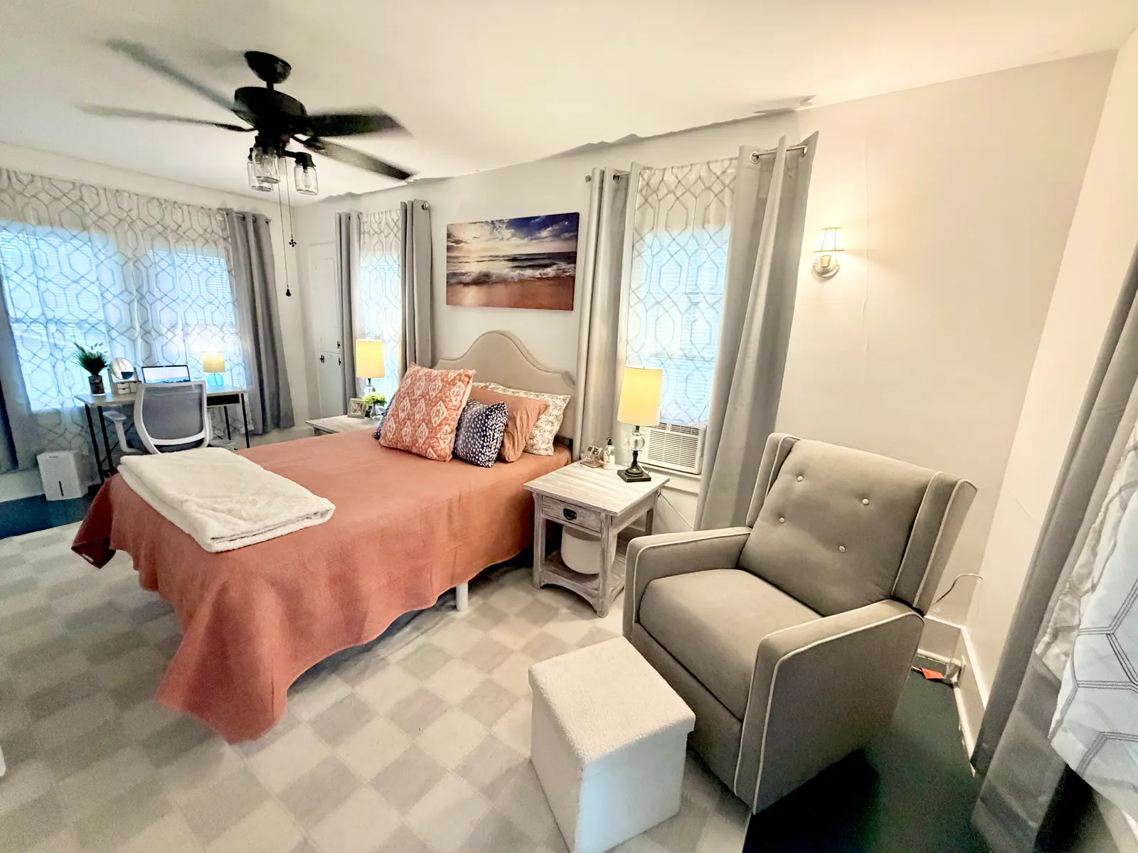 Main bedroom with a queen bed, an upholstered recliner and footstool, two lamplit nightstands, a window air-conditioning unit and a desk in the far corner, on carpet under a ceiling fan