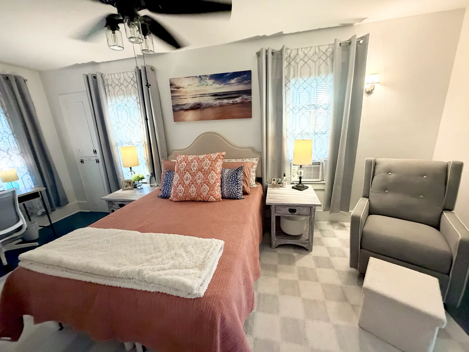 Main bedroom from the other side, with the queen bed between two curtained windows, the recliner beside it and the window air-conditioning unit above the nightstand