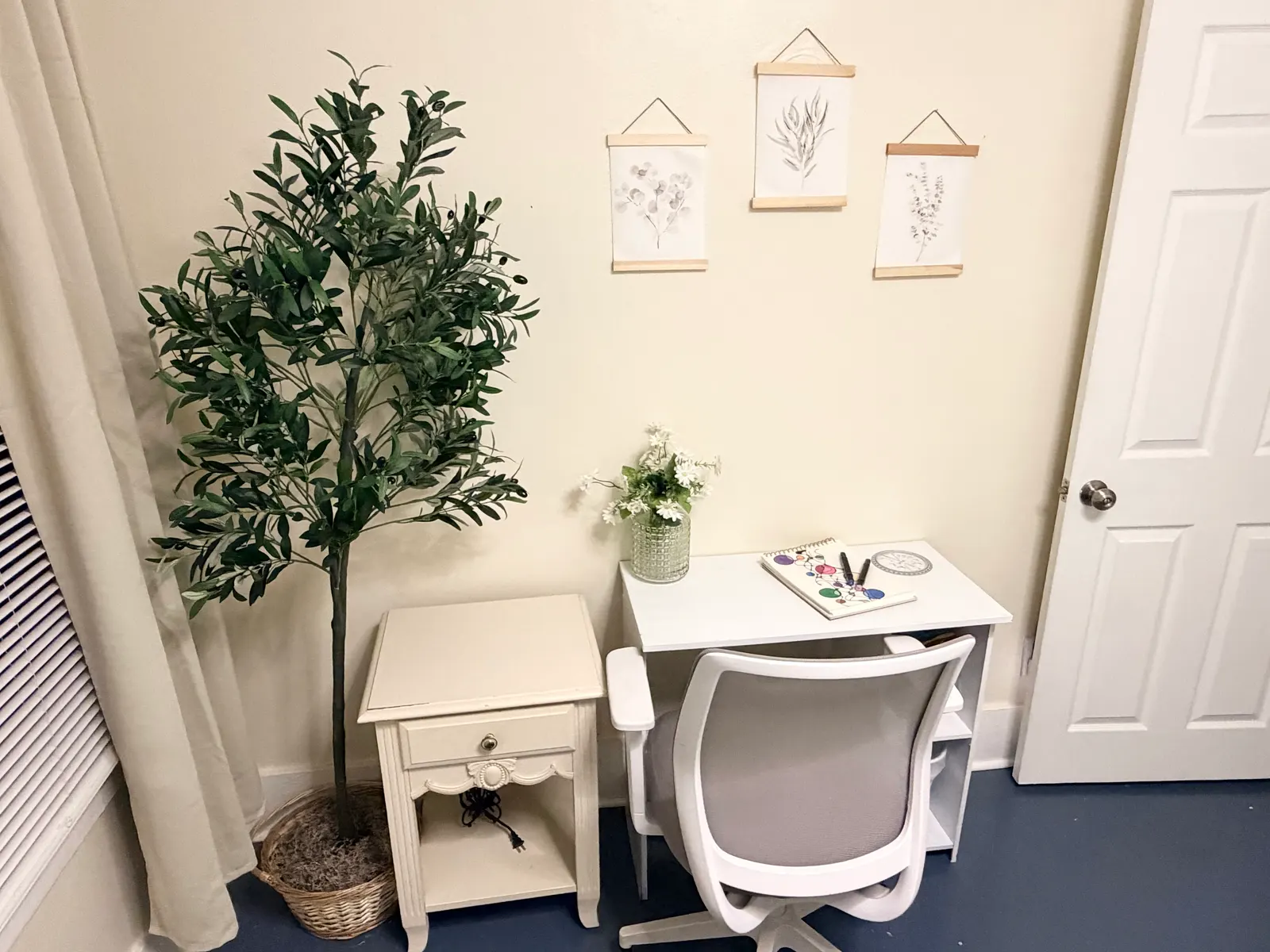 The desk corner of the second bedroom, with an office chair, a small bedside table and a tall potted plant beside the door