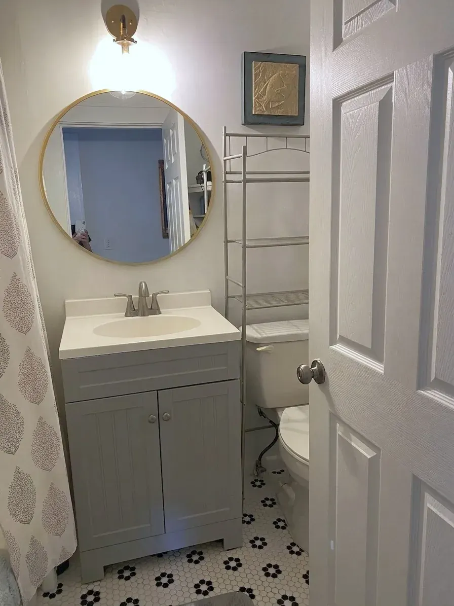 Bathroom with a single-sink vanity, a round mirror, a wall sconce, an over-the-toilet shelving rack and a shower behind a curtain, on hexagonal tile